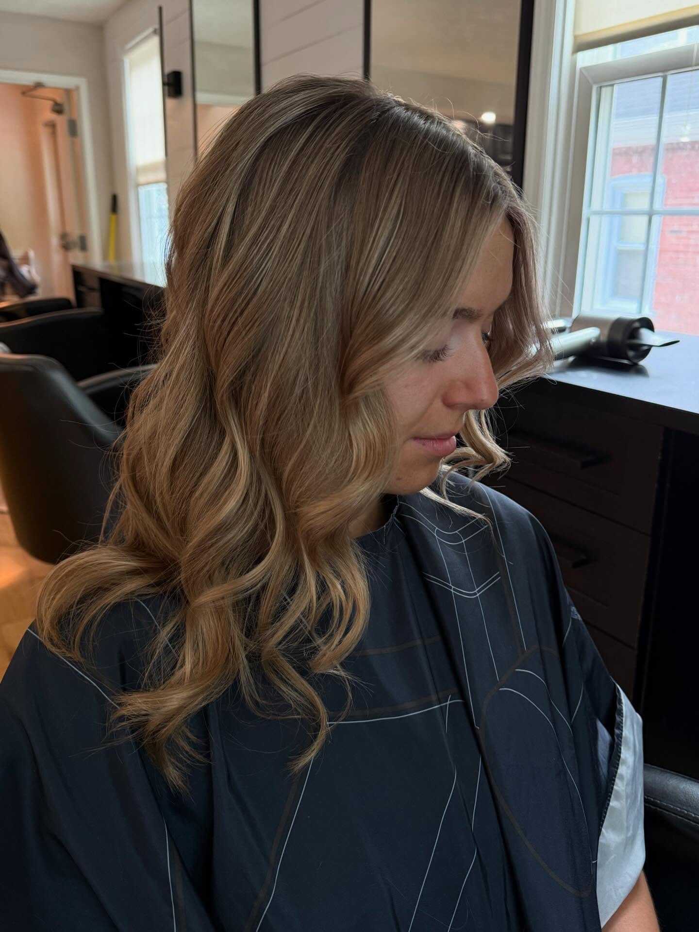 Woman with wavy blonde hair in a salon, wearing a dark cape, looking downward.